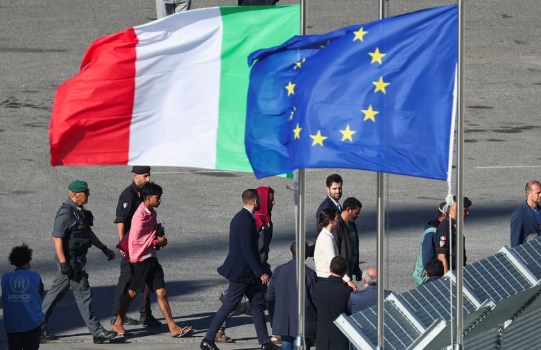 A group of men walk with others in uniform past the flags of Italy and the European Union.