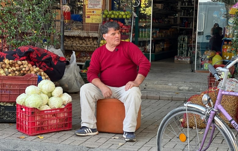 A brown-haired man wearing a red sweater and light pants sits in front of a grocery store.