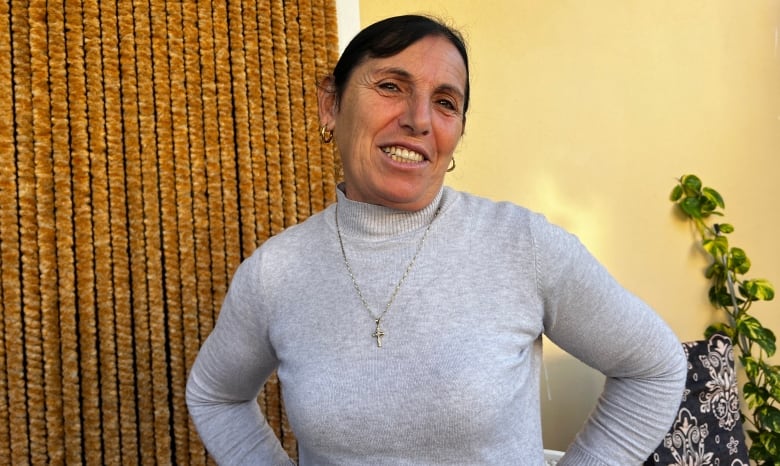 A dark-haired woman wearing a grey turtleneck sweater smiles as she stands in a room.