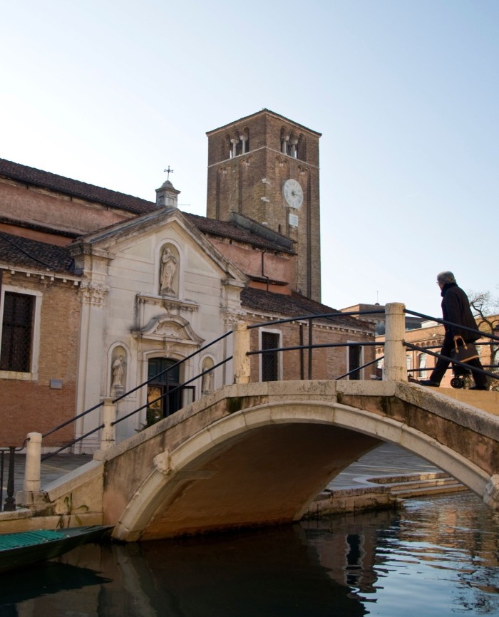 A man walking on a small historic bridge across a narrow canal to the church of San Nicolò dei Mendicoli 