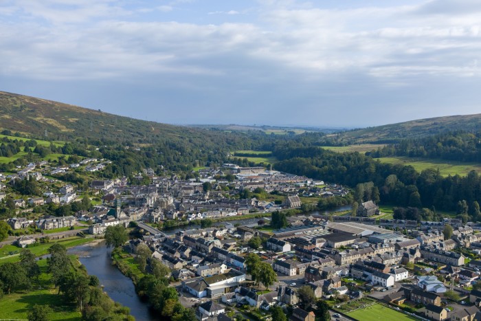 An aerial view of a small town nestled in a valley surrounded by hills and lush greenery. The town is composed of tightly packed stone and modern houses