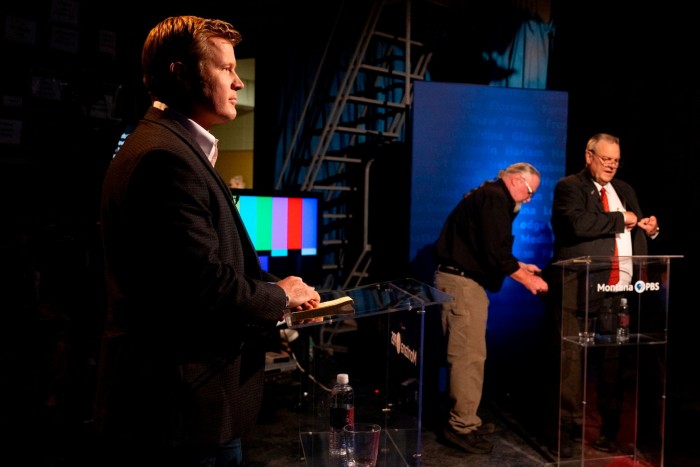 Tim Sheehy, on the left, stands at a debate podium holding a notebook, while Jon Tester, on the right, adjusts his attire at another podium and a technician assists with equipment in the background