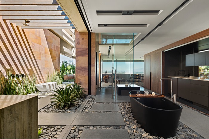 A luxurious modern indoor-outdoor living space featuring a sleek black freestanding bathtub on a bed of pebbles, with a view of a glass-enclosed living area