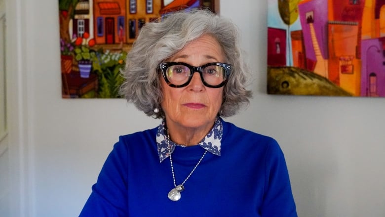 A woman with grey hair and dark glasses wearing a bright blue sweater stands in a hallway in front of two brightly-coloured paintings.