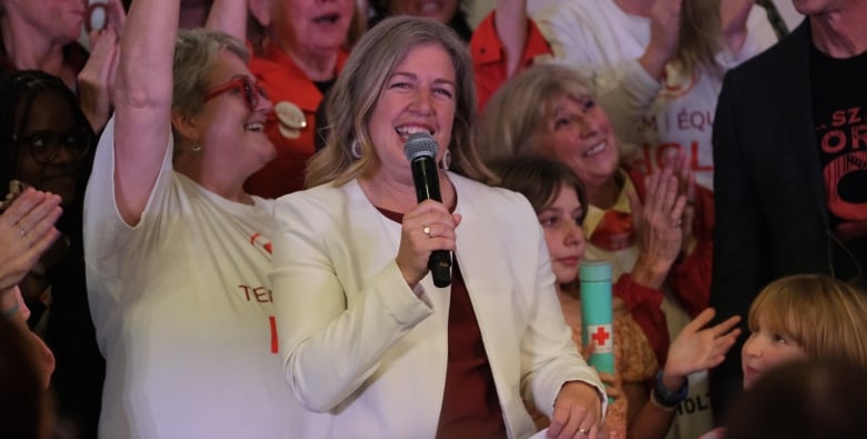 A woman in a white blazer with long blonde hair stands in front of a crowd smiling and holding a microphone.