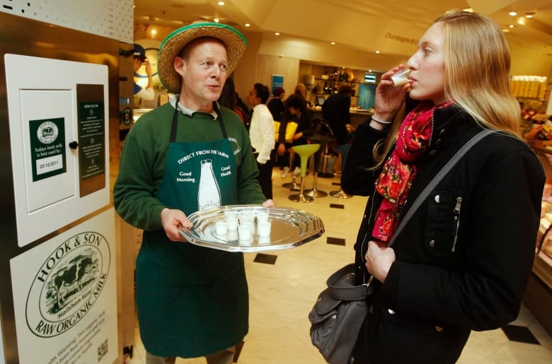 A man dressed as a farmer offers a shopper a sample of milk to drink.