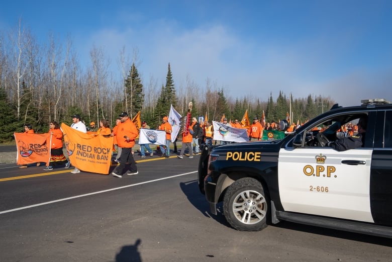 A police cruiser is seen on a highway as a large crowd of people dressed in orange walk by.