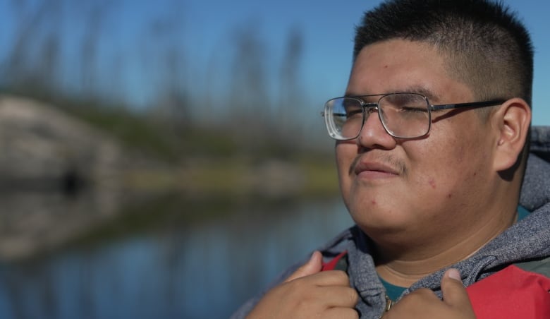 A young man wearing glasses, a hoodie and a life jacket poses for a photo while looking at a river.