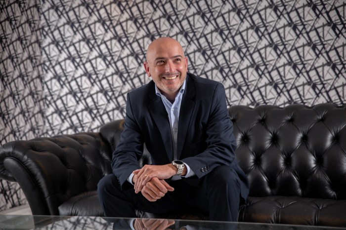 A bald man in a dark suit and light shirt smiles while sitting on a black leather couch