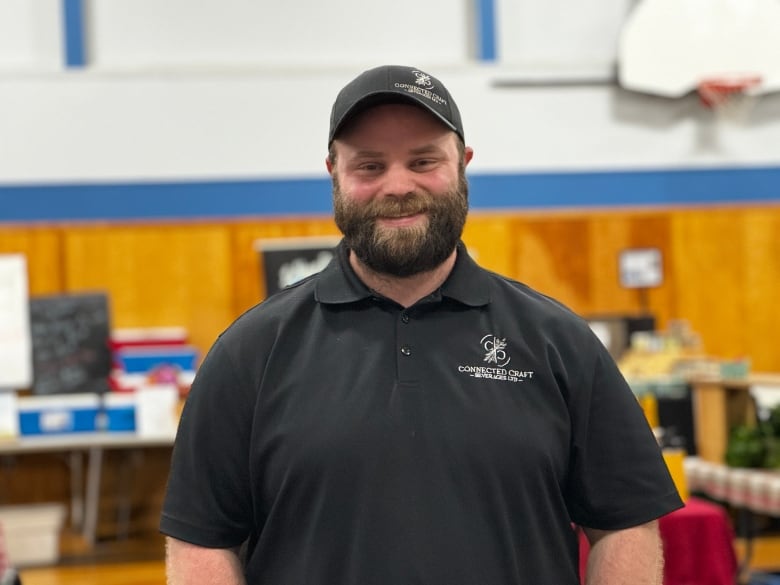 A man wearing a black ball hat and black golf shirt smiles while standing at his booth at the Lunenburg Farmers' Market.