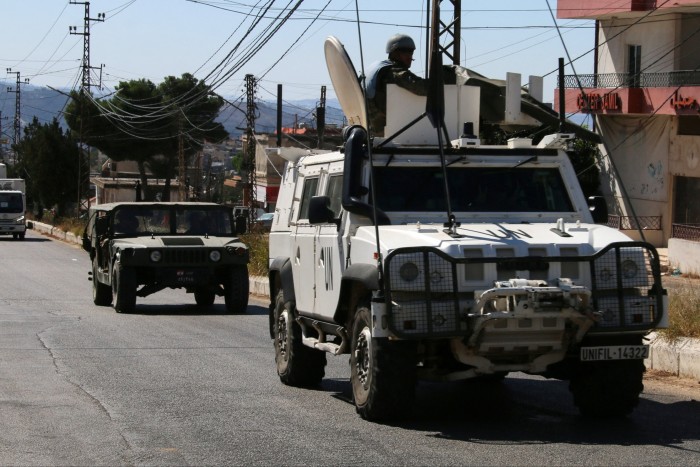 A UN peacekeeper vehicle, marked with &quot;UN&quot; and a Unifil license plate, drives alongside a Lebanese army vehicle in  southern Lebanon. A soldier is visible in the turret of the UN vehicle. Buildings and power lines are seen in the background.