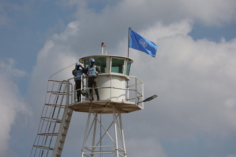 Two UN peacekeepers in blue hats stand on a watchtower flying the blue UN flag. 