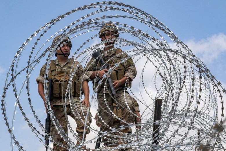 Two men in army fatigues stand behind a barbed wire fence. 