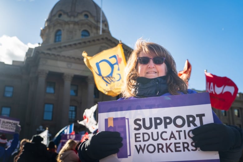 A woman holds a sign that says 'SUPPORT EDUCATION WORKERS' in front of the legislature building. Flags are also visible. 