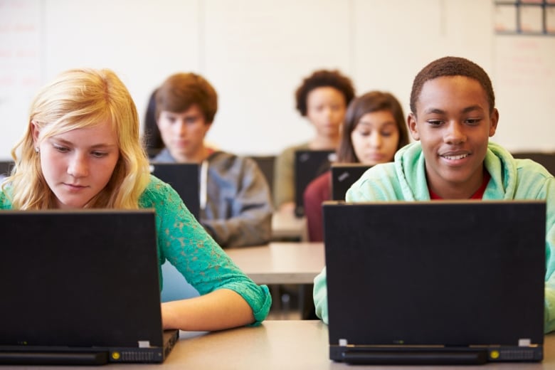 Teenage students in a classroom are seen sitting in rows, looking down at open laptop computers before each of them.