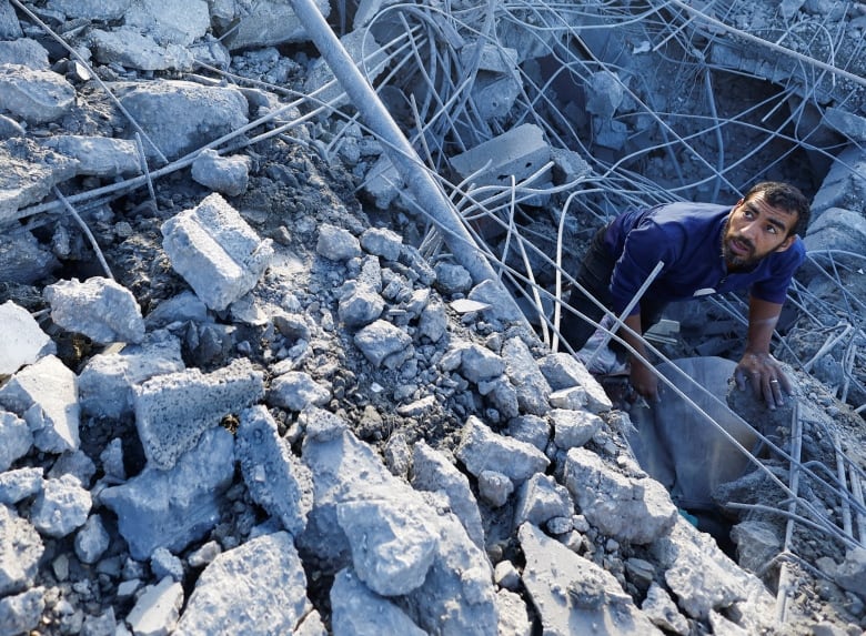 A bearded man in a tshirt is shown crouching, surrounded by rocks and concrete debris.