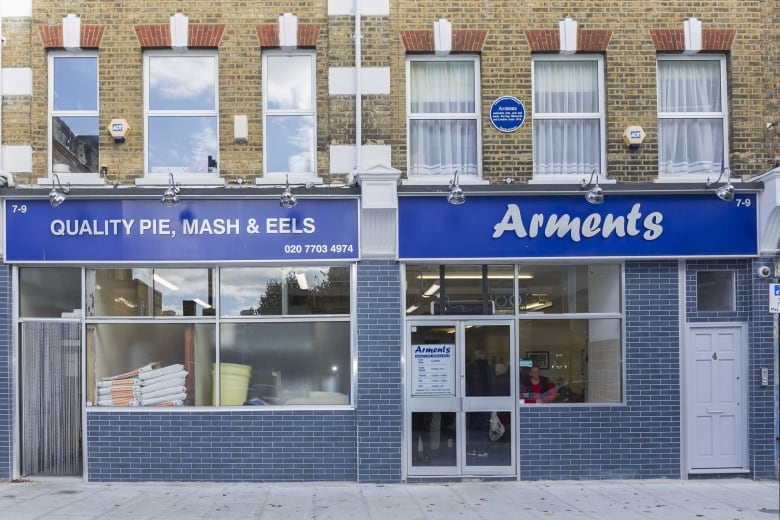 A blue brick storefront with a blue sign that reads "Quality pie, mash & eels" over one store window, and "Arments" over the other window.