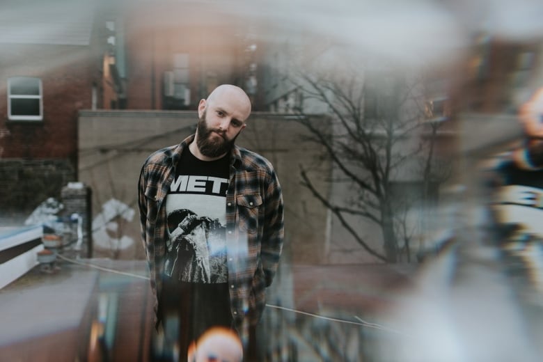 A man stands on a rooftop. He is bald and has facial hair, and is wearing a plaid shirt over a tee shirt that says "METZ."