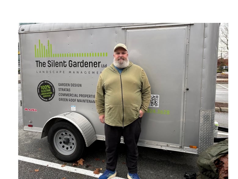 A man with a white beard wearing a white green hoodie and baseball cap stands in front of a cargo carrier that is connected to his pick-up truck. 