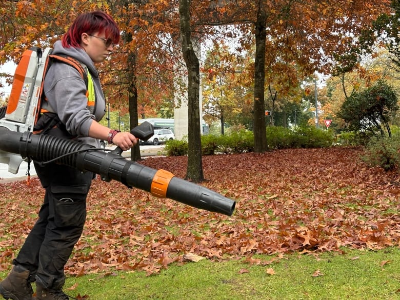 A young woman with short red and dark hair is using an electric leaf blower to blow leaves. 