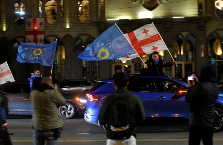 People holding flags lean out of the windows of moving cars as other supporters stand on the street.