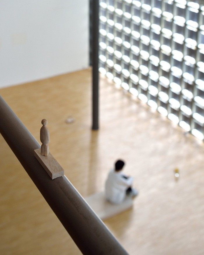 A tiny wooden figurine resting on a handrail overlooking the blurred image of someone sitting on the wooden floor below in ‘Come and Live – Go and Live’ by Rei Naito at Tokyo’s Ginza Maison Hermès Le Forum