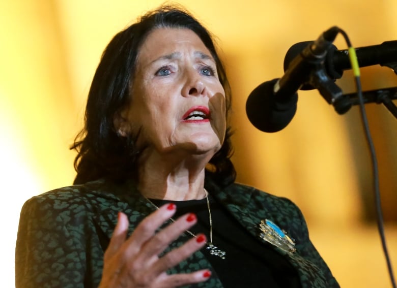 A dark-haired woman, wearing a green and black patterned blazer, speaks into a microphone.