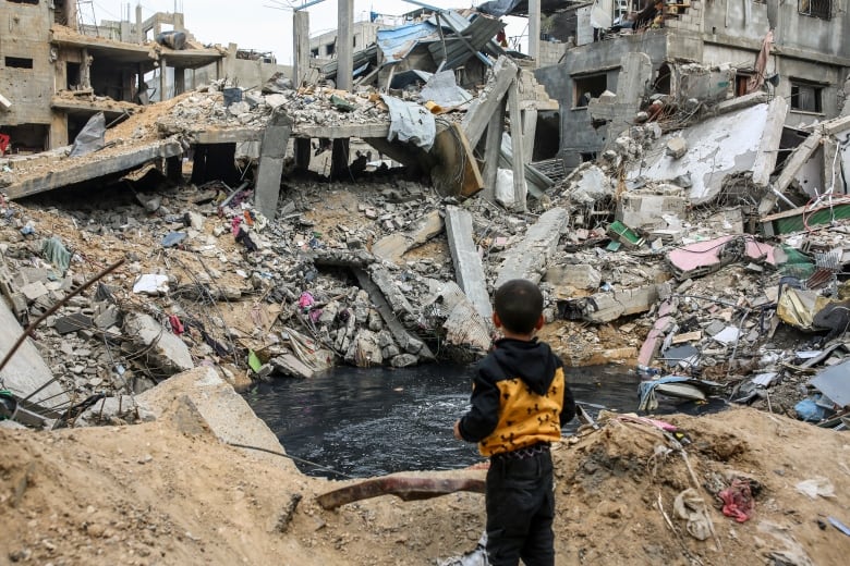 A little boy, pictured from behind, stands in front of a massive pile of rubble.