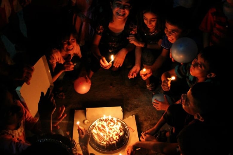 Children sit on the ground in a circle around a birthday cake, their smiling faces lit by the burning candles.