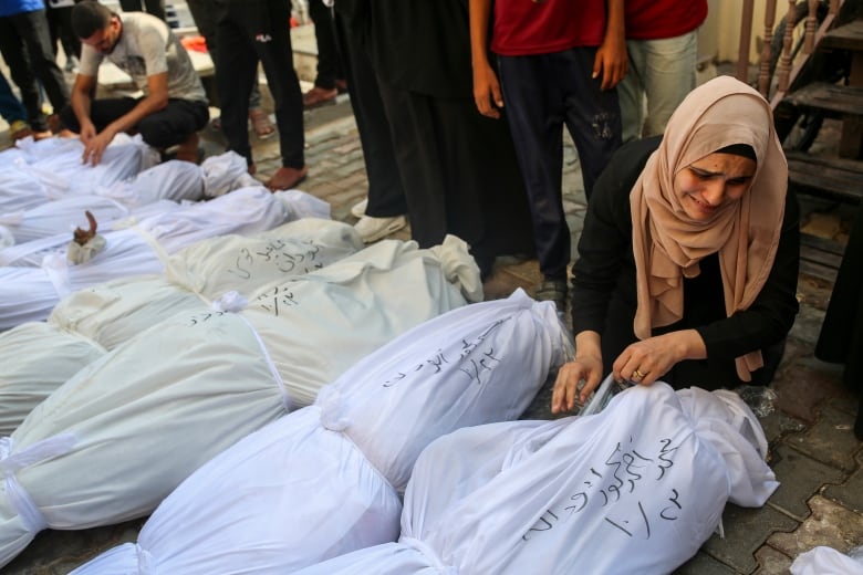 A woman in a headscarf kneels on the ground sobbing next to a row of bodies, wrapped in white cloth, and laid out next to each other. 