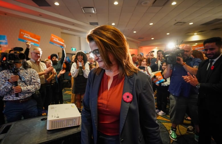 A woman in a blazer and red shirt backgrounded by people with orange signs and clapping