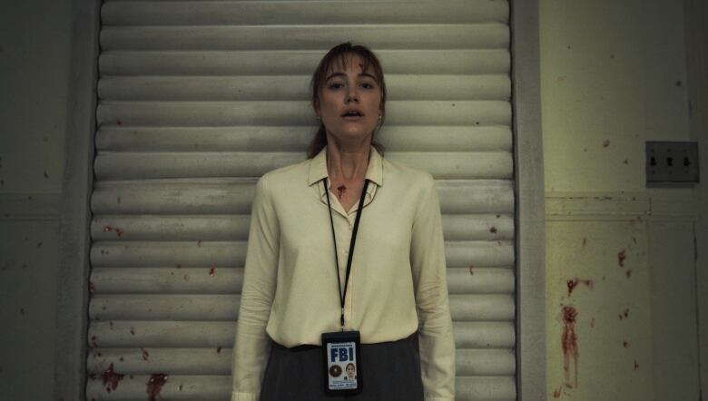 A woman wearing an FBI badge stands against a wall. The wall is spattered in blood and she appears to be terrified. 