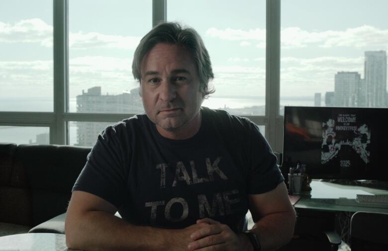 A man in a black t-shirt sits at a desk in front of a window. 