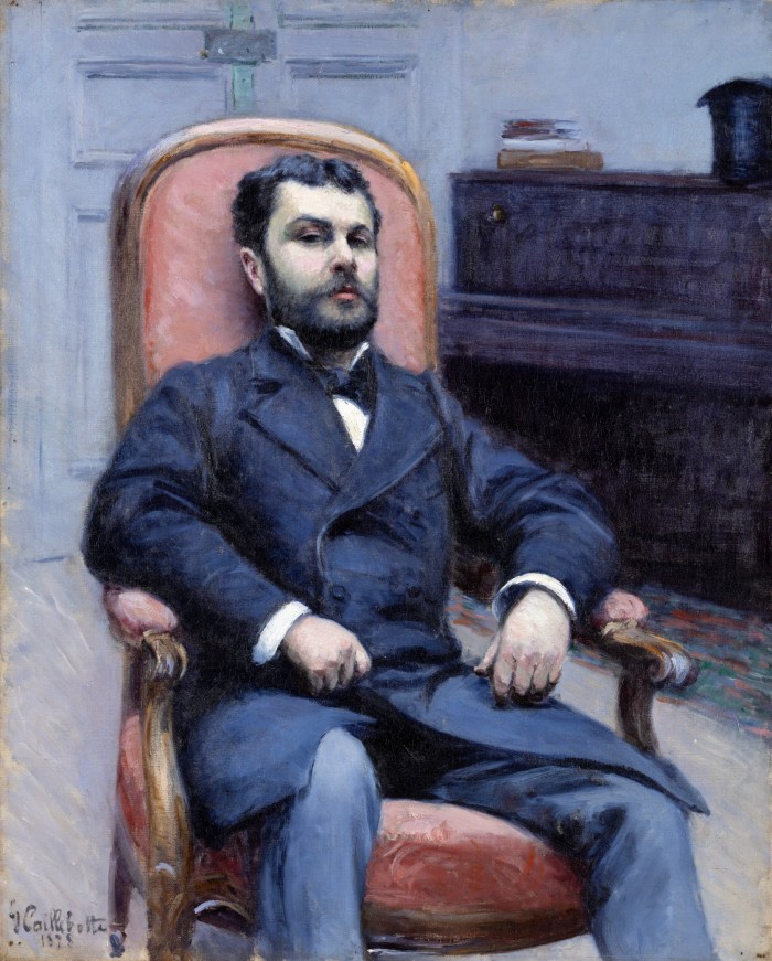 Portrait of a bearded man looking seriously at the viewer, sitting in a pink chair, behind which is a piano and closed wooden doors