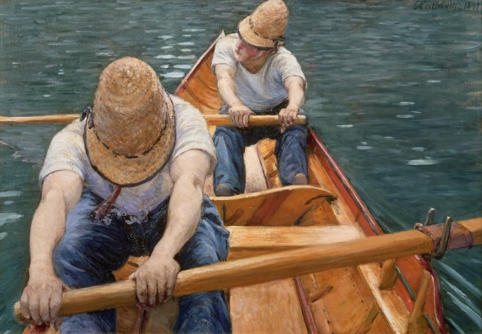 Two men, one in front of the other, strain at the oars while rowing a boat. They wear heavy blue trousers, white T-shirts and straw hats