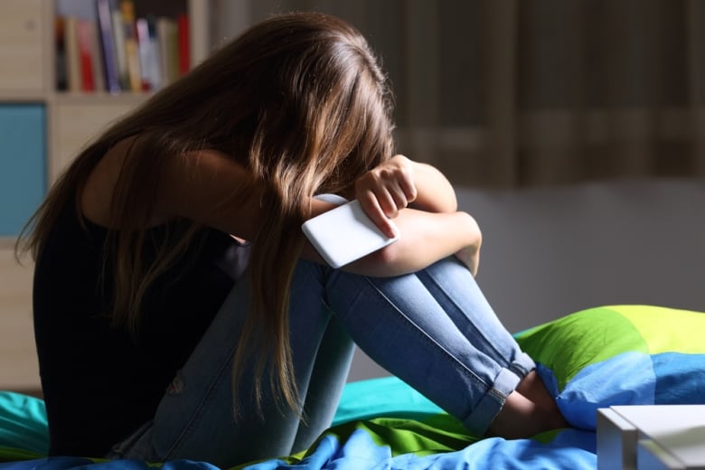 A young girl holds a cellphone in her hands as she puts her head into her knees.