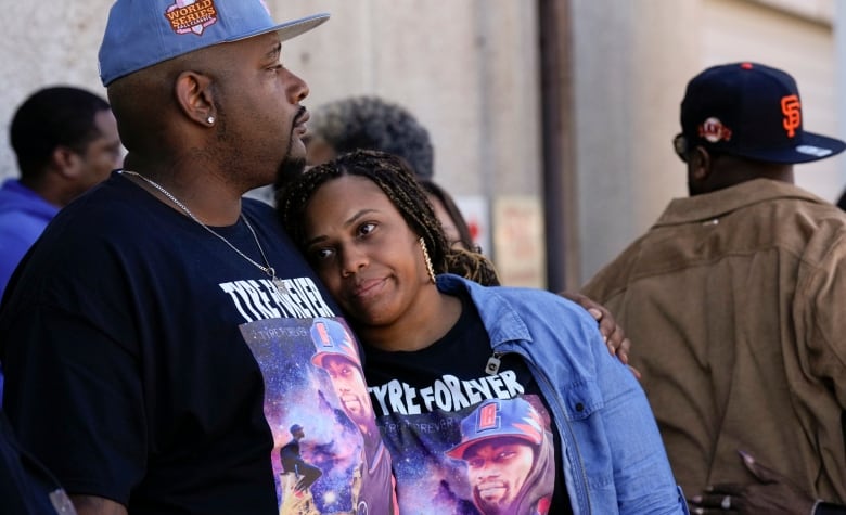 A woman leans against the chest of a man as he gazes off into the distance. He has an arm around her and they are both wearing matching shirts that say "Tyre Forever". 