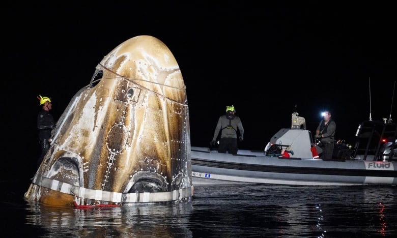 A brown space capsule floats on the sea in the dark, as a boat approaches