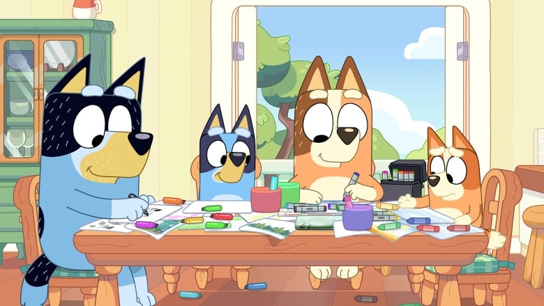 A screenshot from the animated show Bluey shows four cartoon dogs, two parents and two kids, sitting at a table and making drawings. The table is covered in art supplies and sheets of paper. 