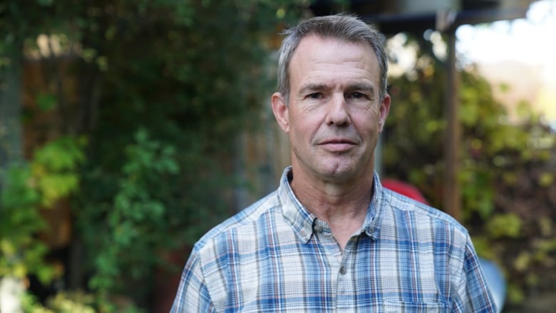 A man stands in a plaid shirt in his lawn not smiling in his backyard.