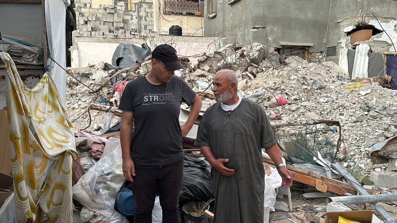 Two men stand in front of rubble. 