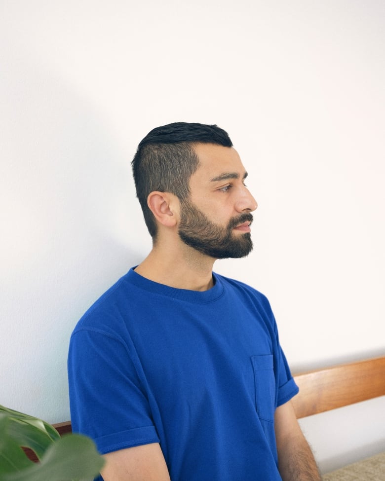 A medium-length portrait of a man with short dark hair and tightly cropped beard, wearing a blue shirt and sitting in profile. 