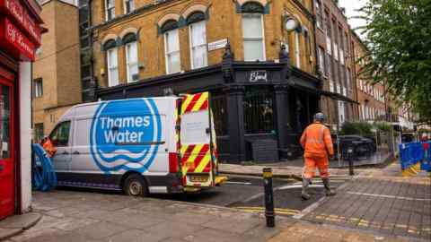 A maintenance engineer in an orange high-visibility suit and helmet walks towards an incident site in London. A Thames Water van is parked nearby with its rear doors open, and blue hoses are draped over the side.