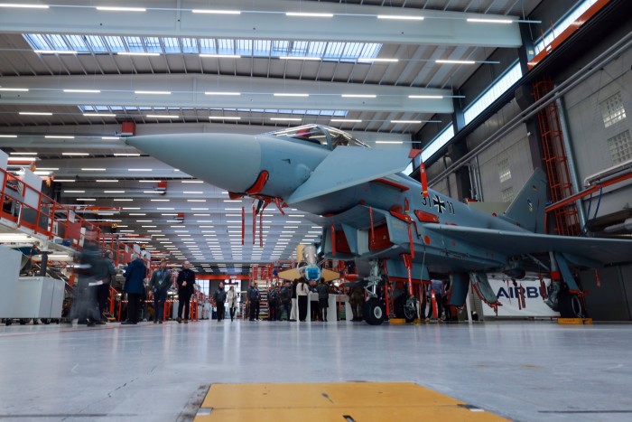 A Eurofighter Typhoon is showcased inside an Airbus manufacturing plant, with people gathered around in the background. The aircraft is surrounded by equipment and appears in a large, well-lit industrial space.