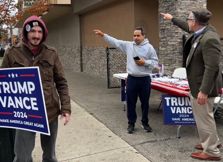 People point, near Trump Vance signs on a sidewalk stand.