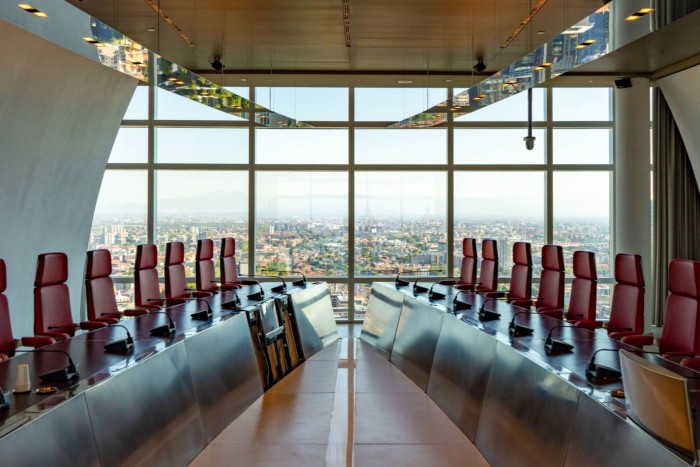 A modern boardroom featuring a long table with red chairs and microphones, set against a large window offering a panoramic view of the city.