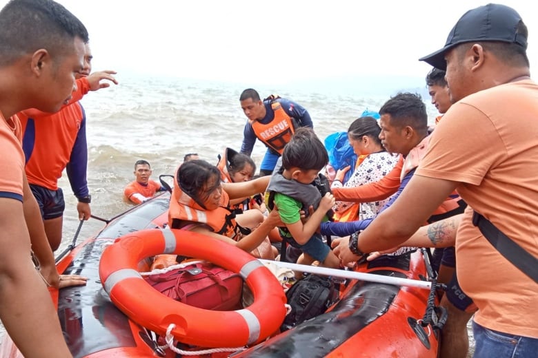 Several people, many of them small children, wear life jacket vests as they're in a rubber beat on water near a shore.