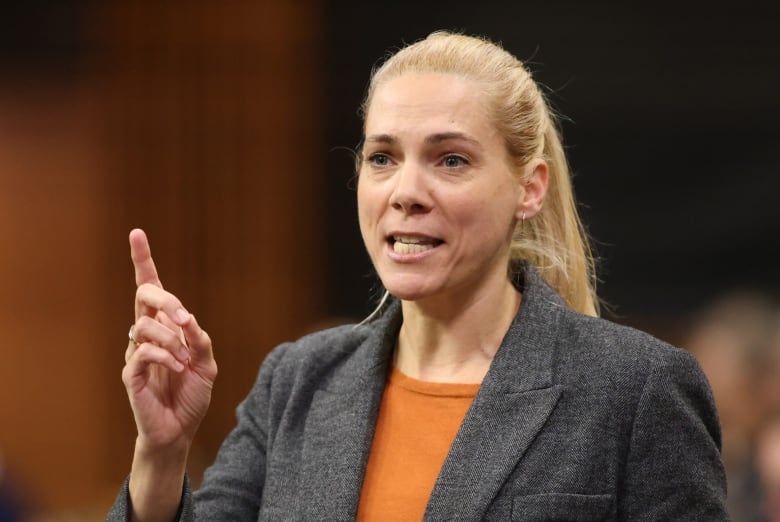 A woman wearing a shirt and an orange blazer is photographed mid-speech. 