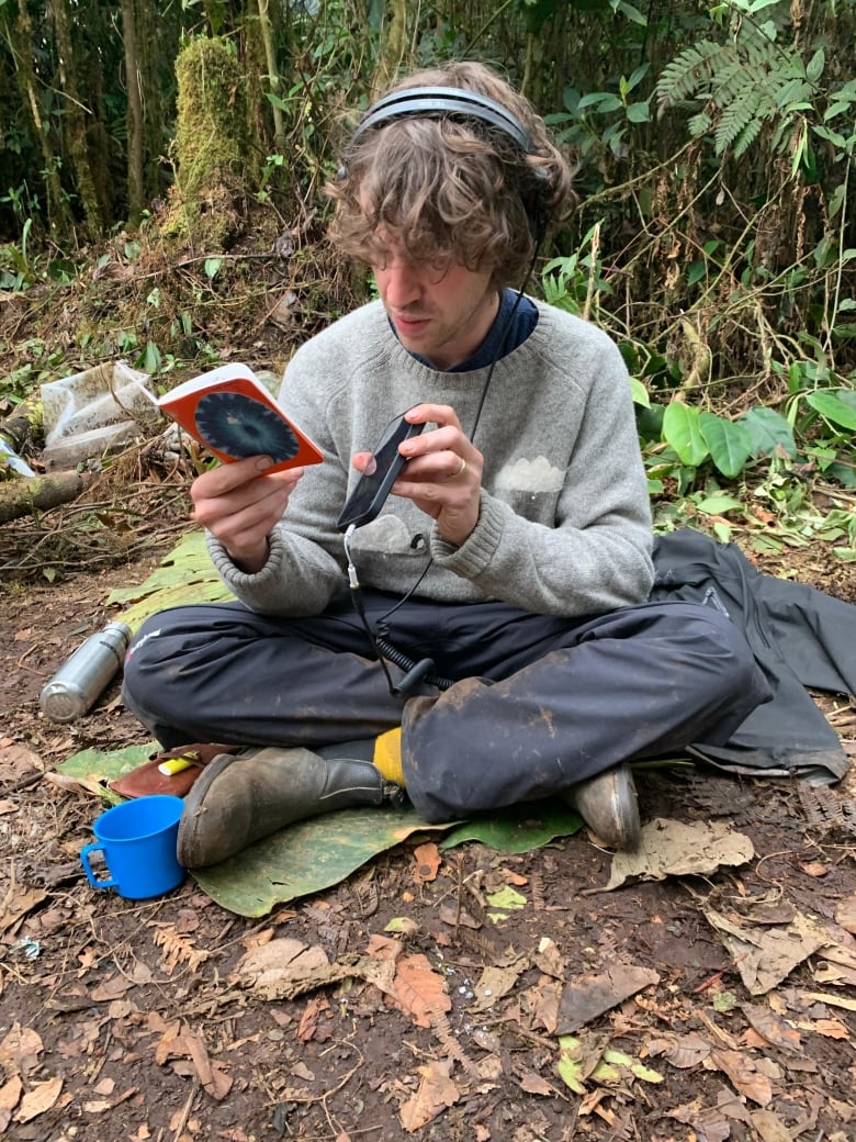 A man with shaggy, lightly curled hair sits on the forest floor with his legs crossed, holding a notebook in one hand while listening to headphones attached to a mobile phone. 