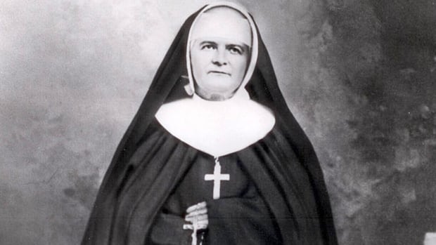 A black and white image of a woman in a black nun's habit, with white trim, and a black gown, wearing a cross around her neck.